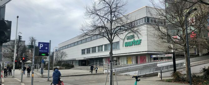 Ein E Scooter steht im Vordergrund. Er ist grün. Dahinter ist die alte Galeria Kaufhof Filiale in Cottbus zu sehen.