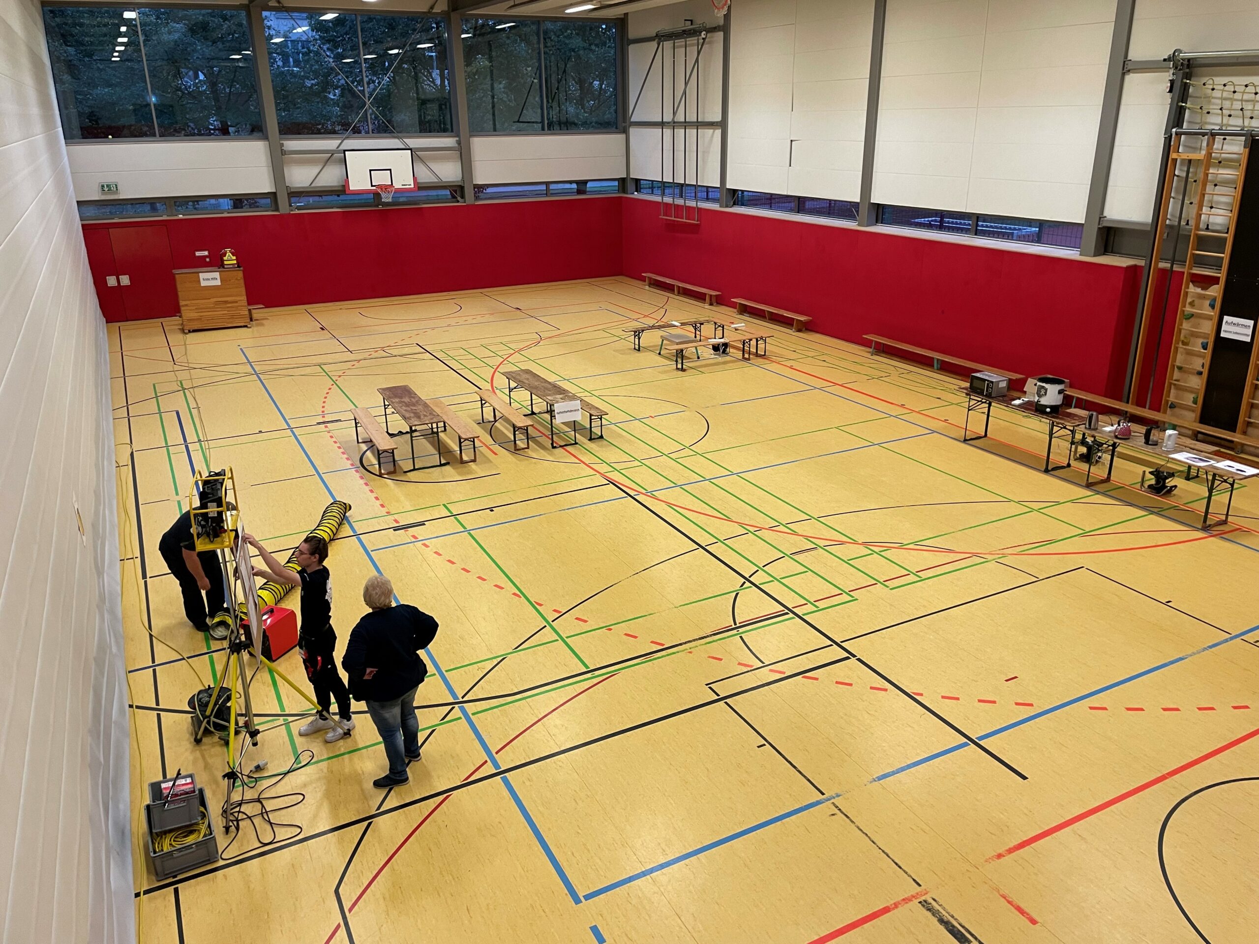 In einer Turnhalle stehen Bänke. Menschen bauen eine Notfallstation mit einer Küche auf.