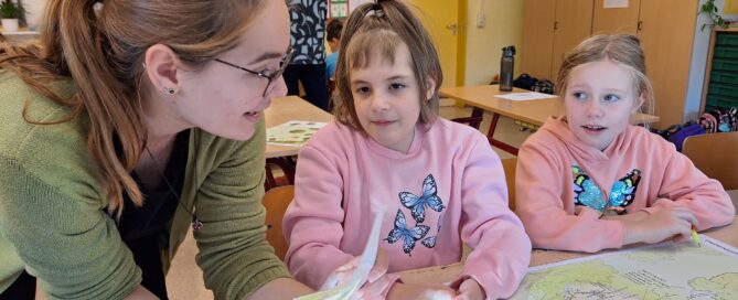 Zwei Kinder sitzen in einem Raum. Eine Frau mit Brille, welche die Erzieherin ist, erklärt ihnen etwas.