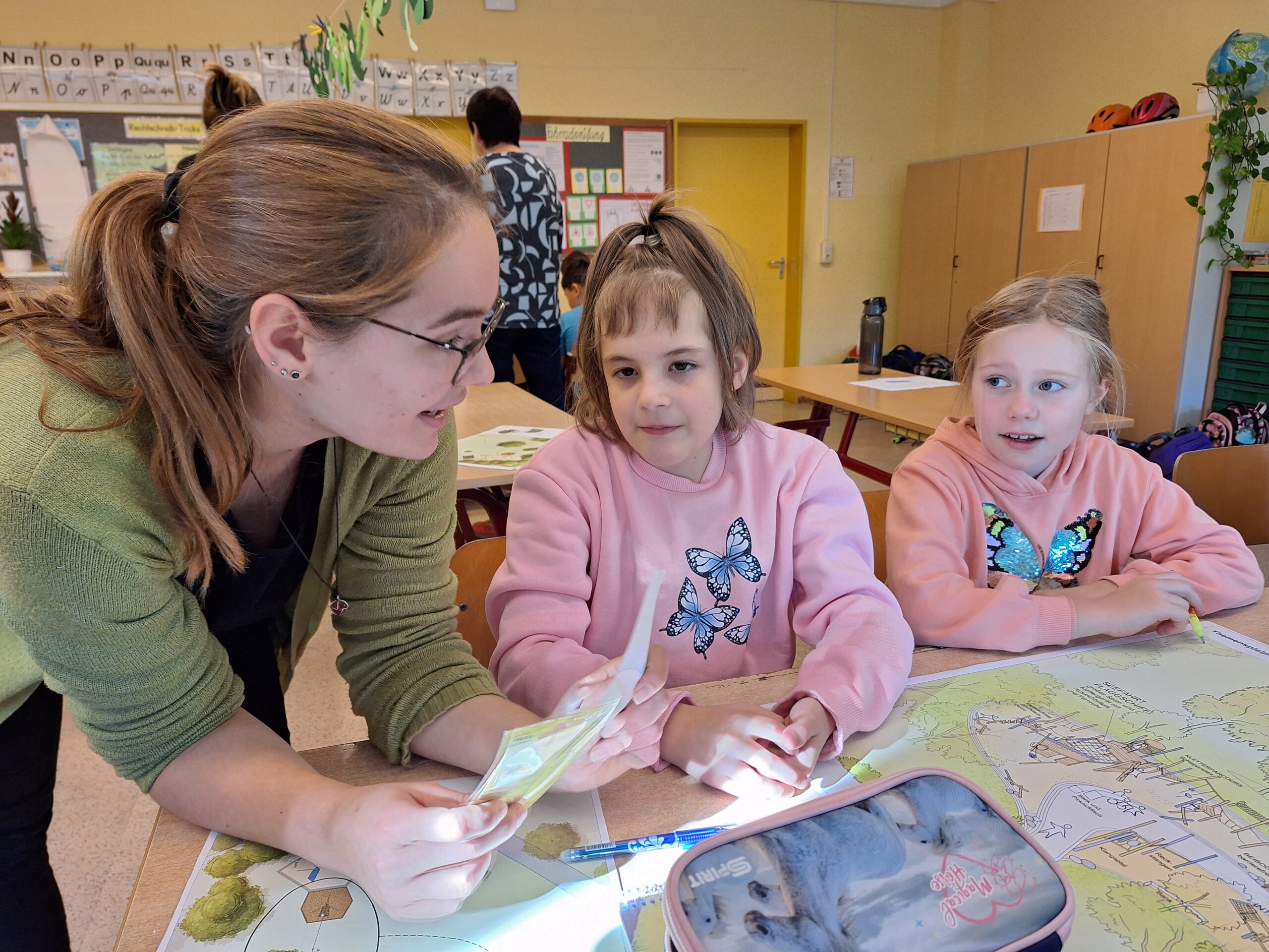 Zwei Kinder sitzen in einem Raum. Eine Frau mit Brille, welche die Erzieherin ist, erklärt ihnen etwas.