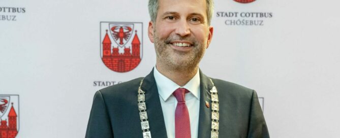 Tobias Schick als Oberbürgermeister mit Anzug und einer roten Krawatte steht vor einer Wand mit dem Logo der Stadt Cottbus