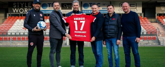 Auf dem Foto sind Vertreter des FC Energie Cottbus und der Koalick Unternehmensgruppe im Stadion zu sehen. Sie halten ein Trikot mit dem neuen Rückenaufdruck in die Höhe.