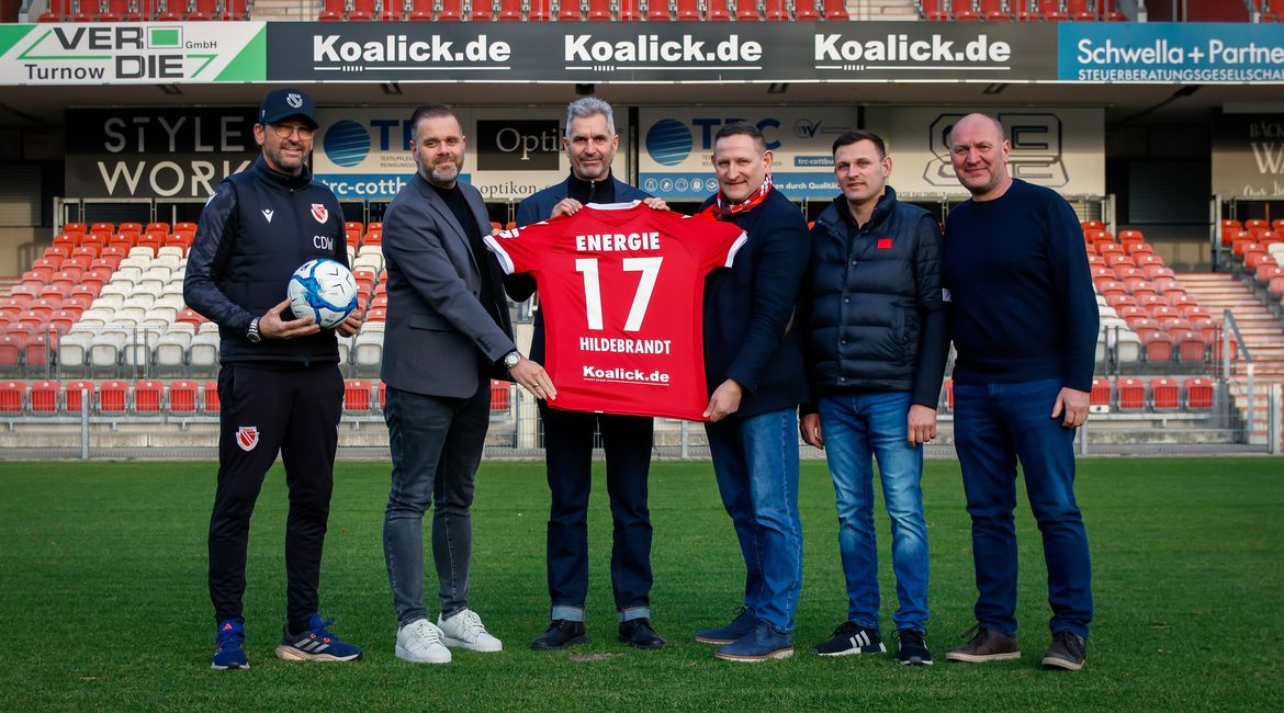 Auf dem Foto sind Vertreter des FC Energie Cottbus und der Koalick Unternehmensgruppe im Stadion zu sehen. Sie halten ein Trikot mit dem neuen Rückenaufdruck in die Höhe.