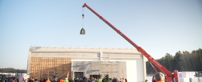 Das Foto zeigt den Rohbau der neuen BIFI-Fabrik. Ein Richtkranz hängt an einem Kran. Vor der Fabrik stehen Menschen.