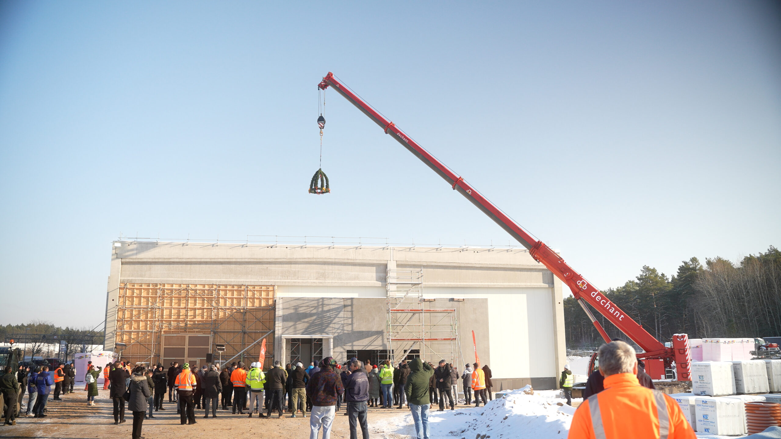 Das Foto zeigt den Rohbau der neuen BIFI-Fabrik. Ein Richtkranz hängt an einem Kran. Vor der Fabrik stehen Menschen.