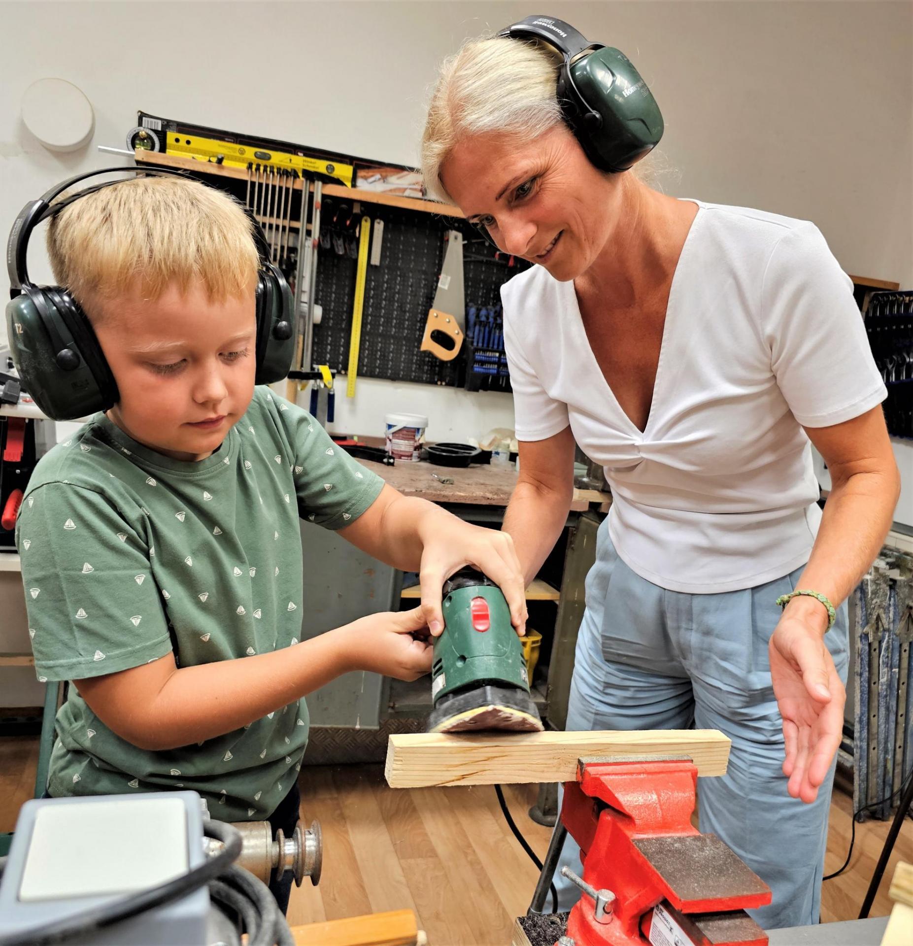 Das Foto zeigt ein Kind mit Gehörschutz, das ein Stück Holz abschleift. Eine erwachsene Frau leitet den Jungen dabei an.