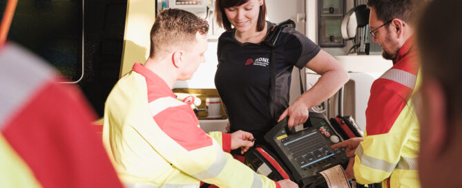 Das Foto zeigt ein technisches Gerät, das beim Rettungsdienst zum Einsatz kommt. Außerdem sind vier Mitarbeitende zu sehen.