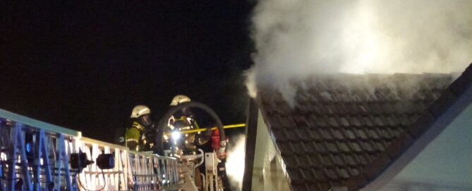 Haus bei dem das Dach qualmt. Außerdem Feuerwehrfahrzeug am Straßenrand.