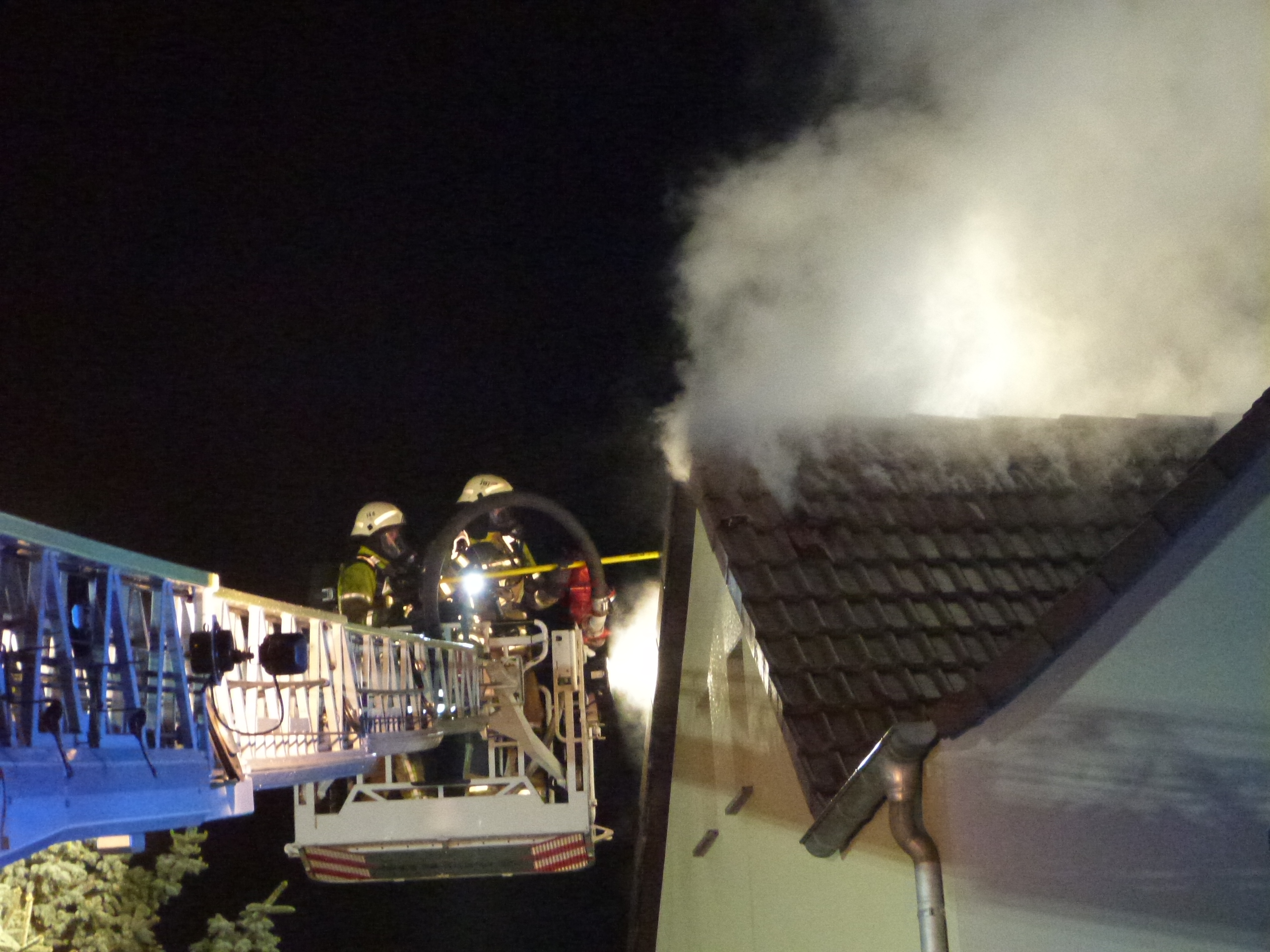 Haus bei dem das Dach qualmt. Außerdem Feuerwehrfahrzeug am Straßenrand.