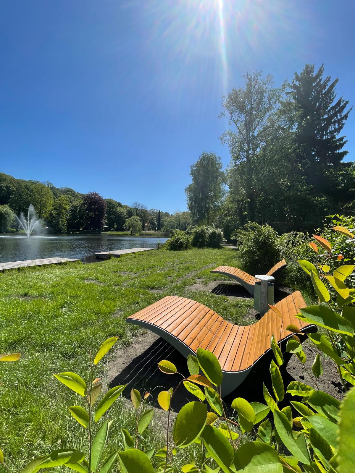 schwanenteich hochkant Das Foto zeigt mehrere Holzliegen am Ufer des Schwanenteichs in Spremberg. Eine Fontäne sprudelt.