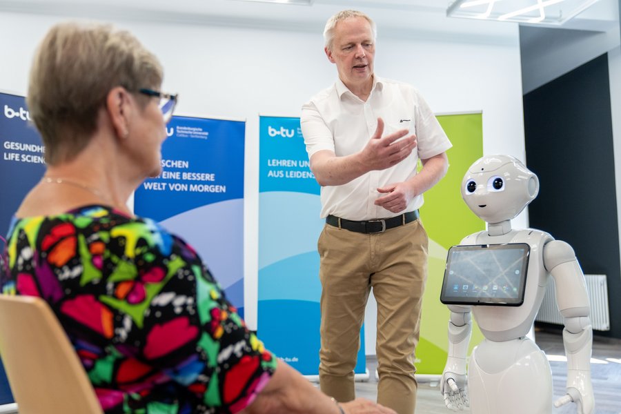 Das Foto zeigt den Roboter Pepper mit einer älteren Dame und einem Herrn im weißen Hemd.