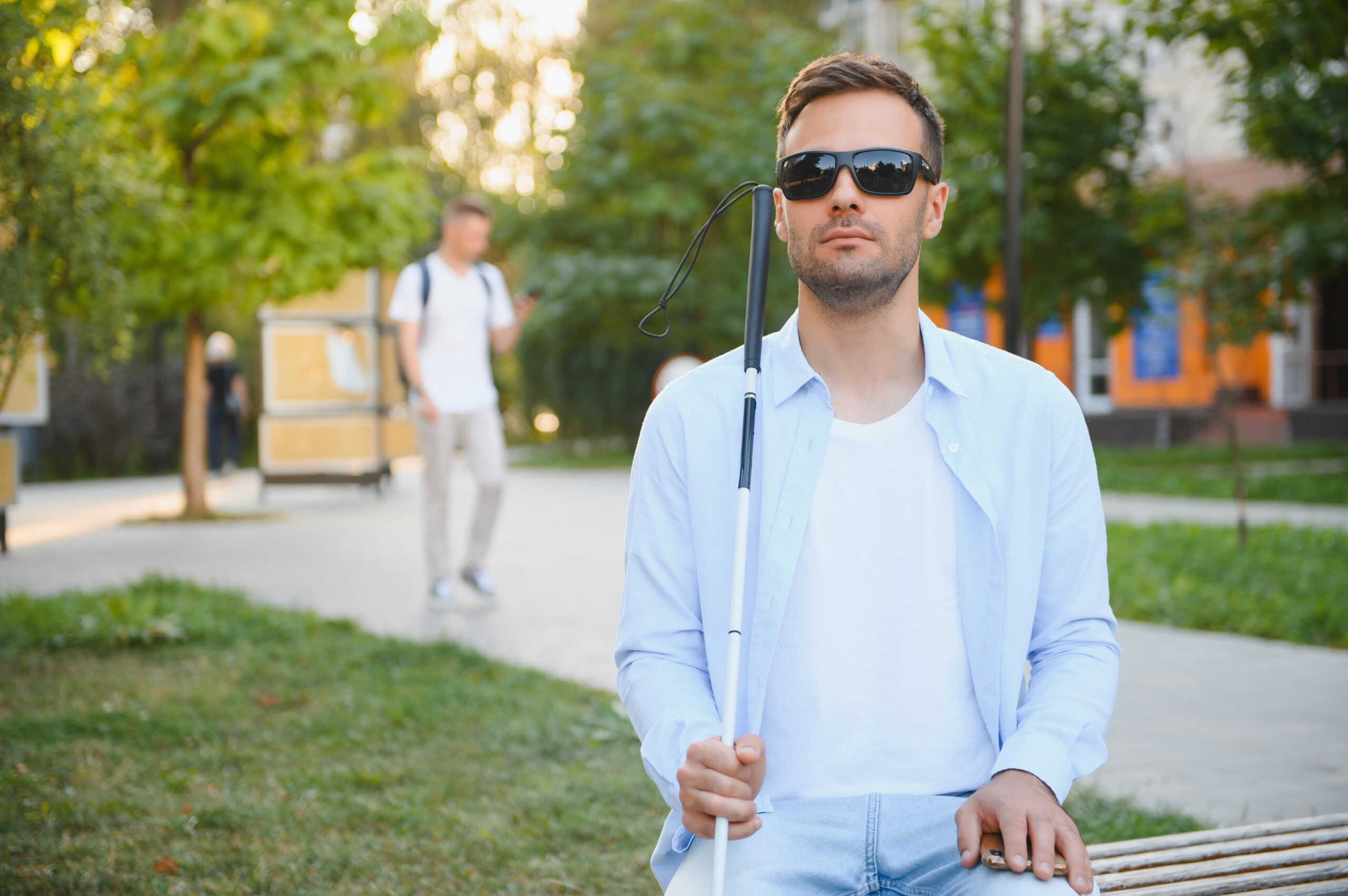 Blind man with a walking stick sitting on a bench at a park Ein blinder Mann mit Sonnenbrille und Blindenstock sitzt auf einer Parkbank.
