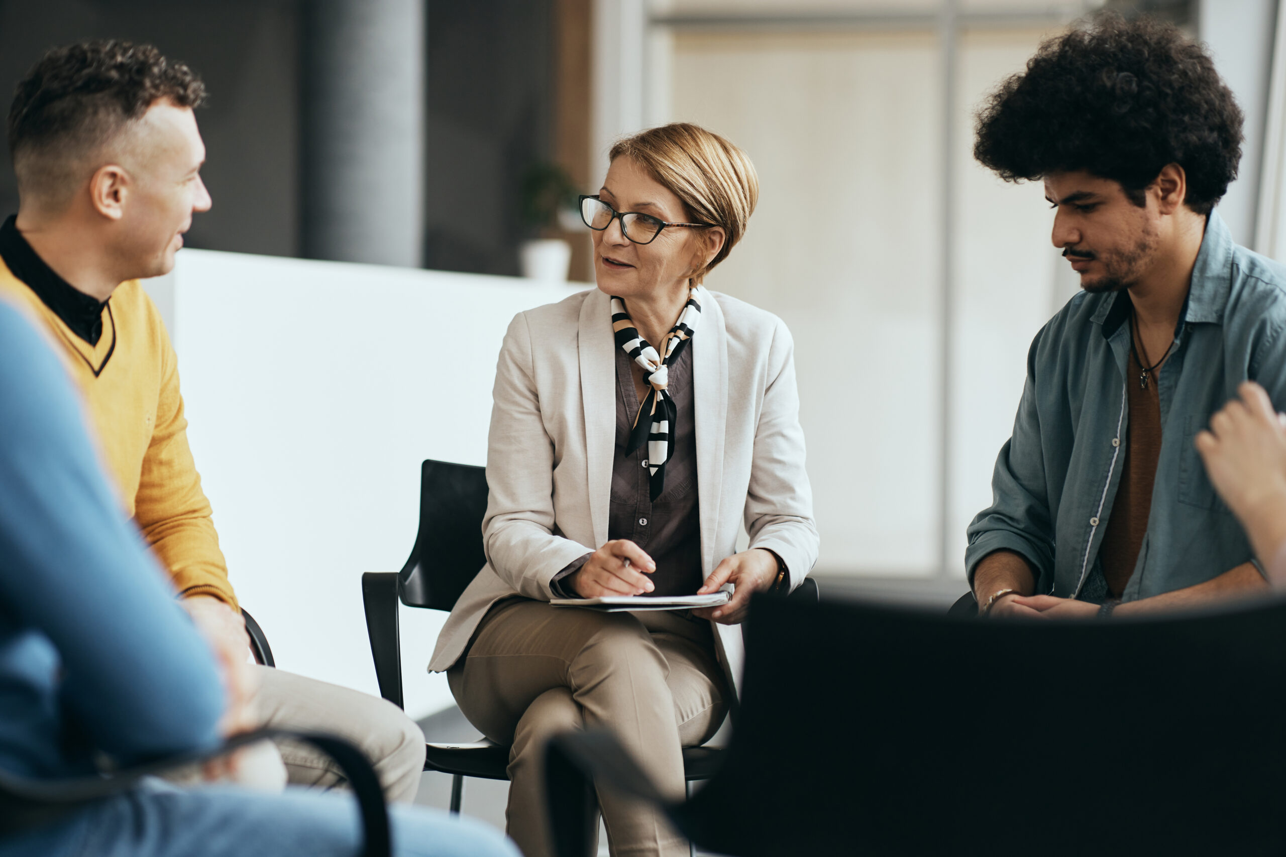 Mature therapist talking to participants of group therapy at mental health center. Eine Therapeutin spricht mit zwei Teilnehmenden einer Gruppe.