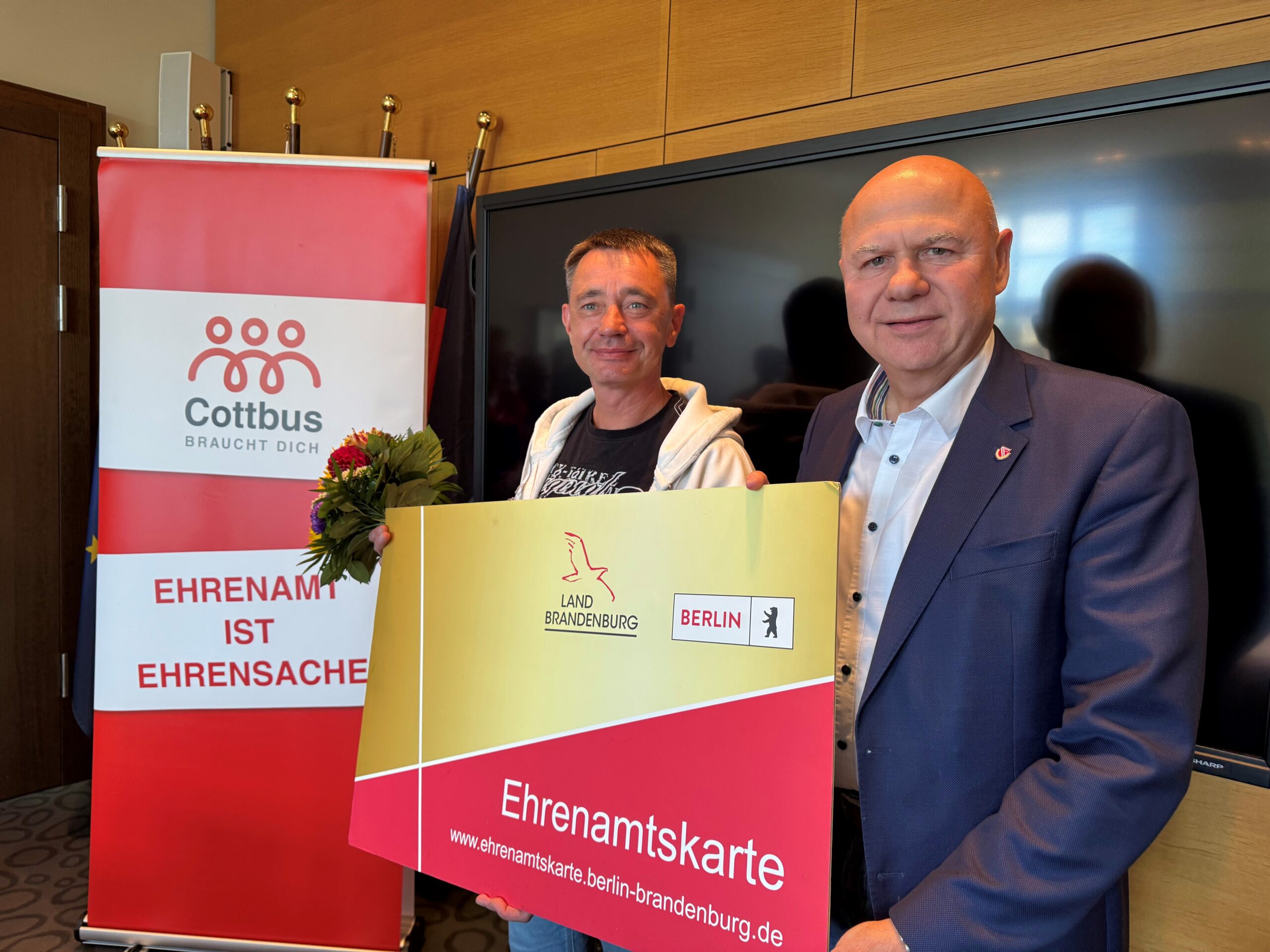 PM_Ehrenamtskarte_Grawunder_Bergner Zwei Männer stehen in einem Raum und halten eine Karte hoch