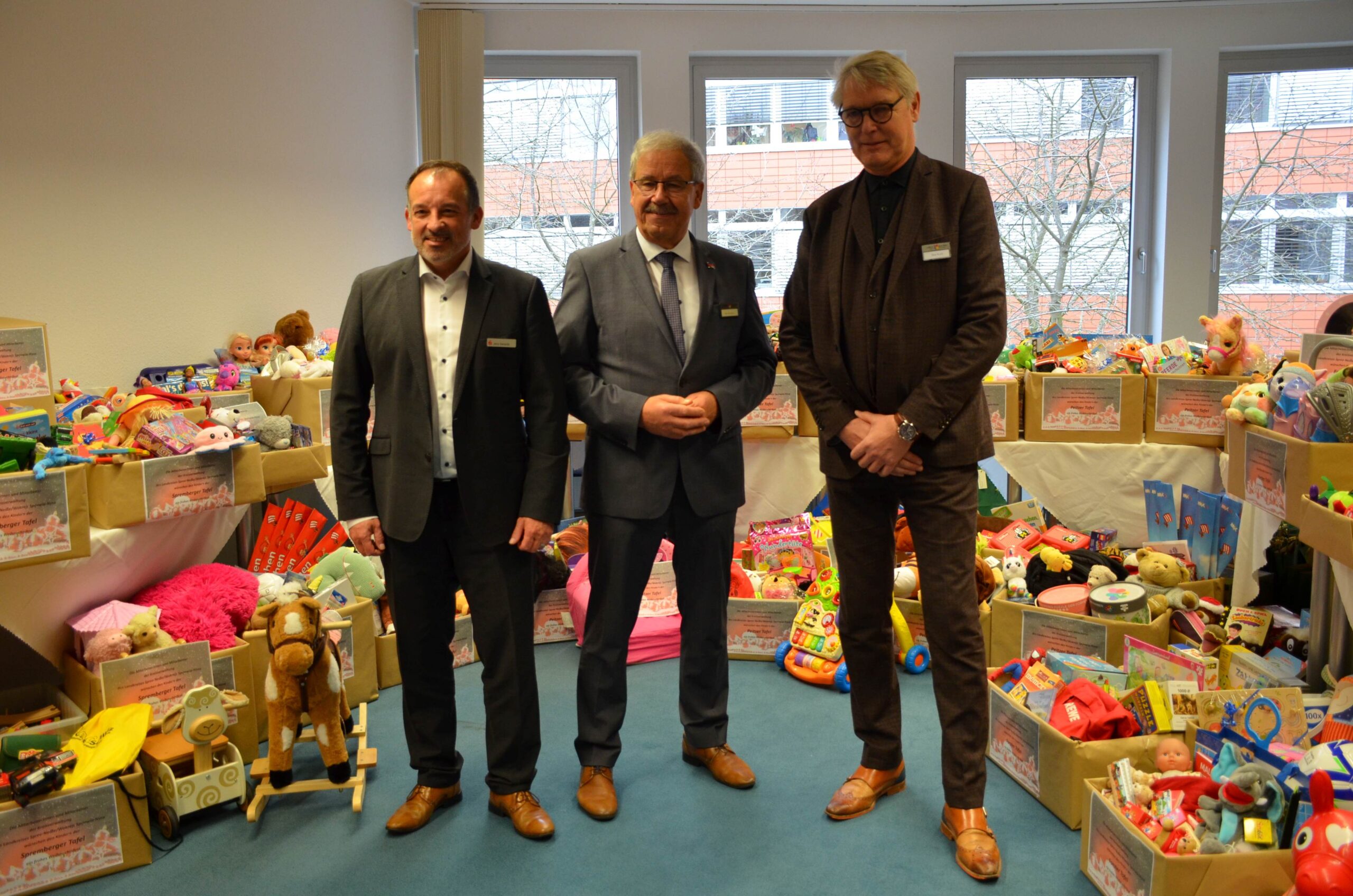 Drei Männer stehen nebeneinander. Im Hintergrund sind Geschenke zu sehen.
