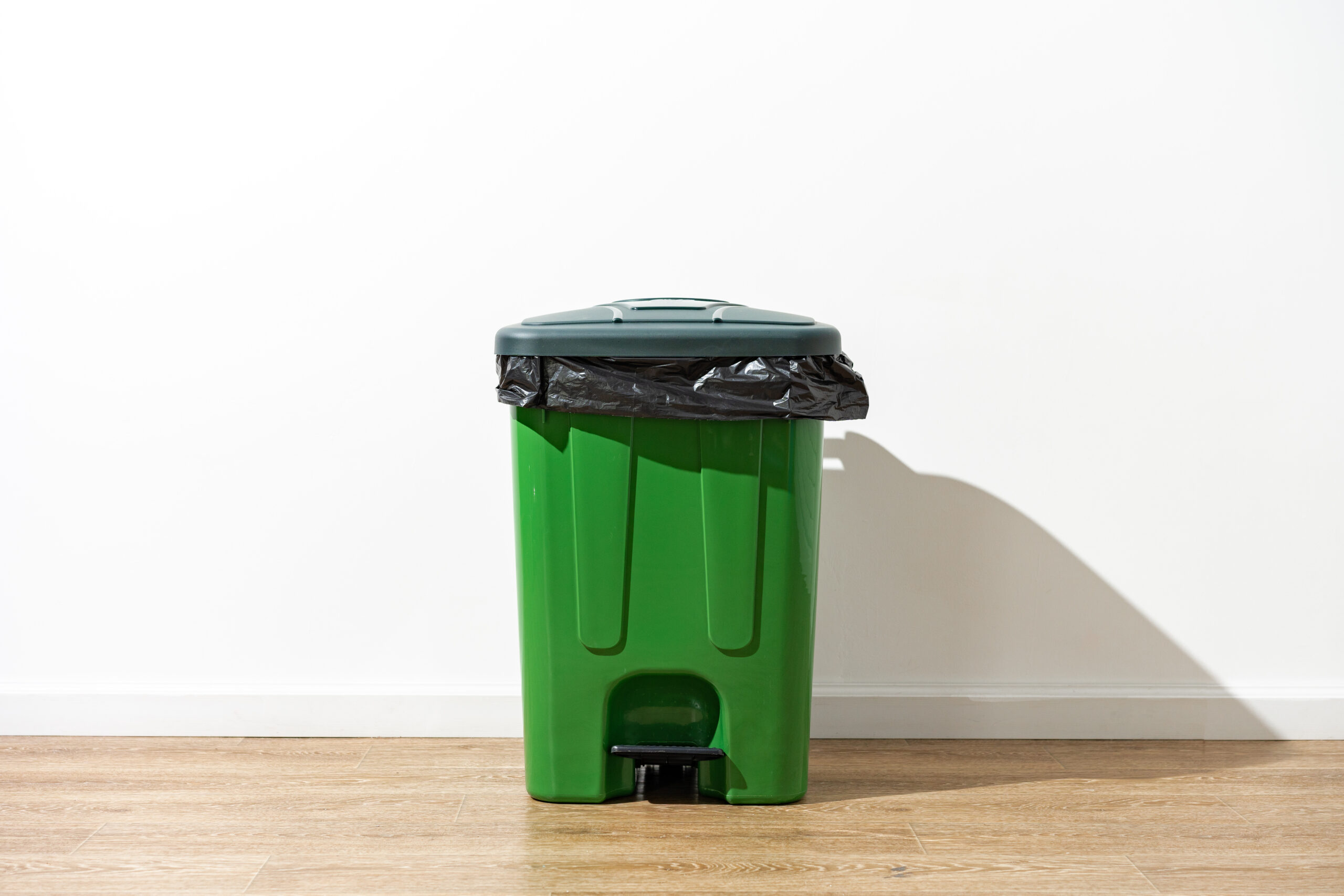 Das Symbolbild zeigt eine grüne Mülltonne vor einer weißen Wand in einem Raum.