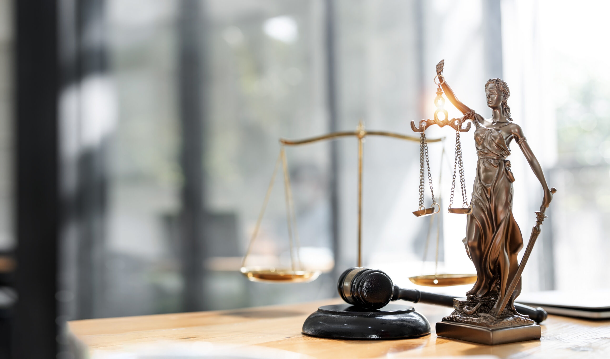 Das Symbolbild zeigt einen Schreibtisch mit einer Statue von Justitia, einer Waage und einem Richterhammer.