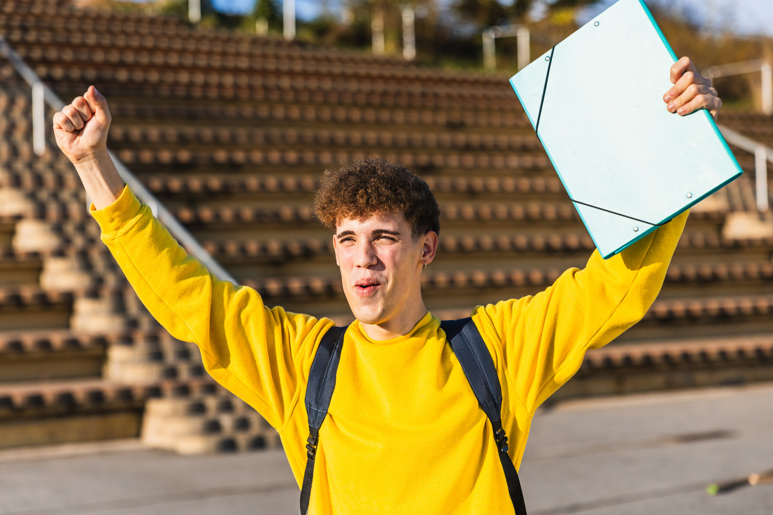 Excited college student celebrating success with raised arms and folder Ein Schüler hält die Arme nach oben