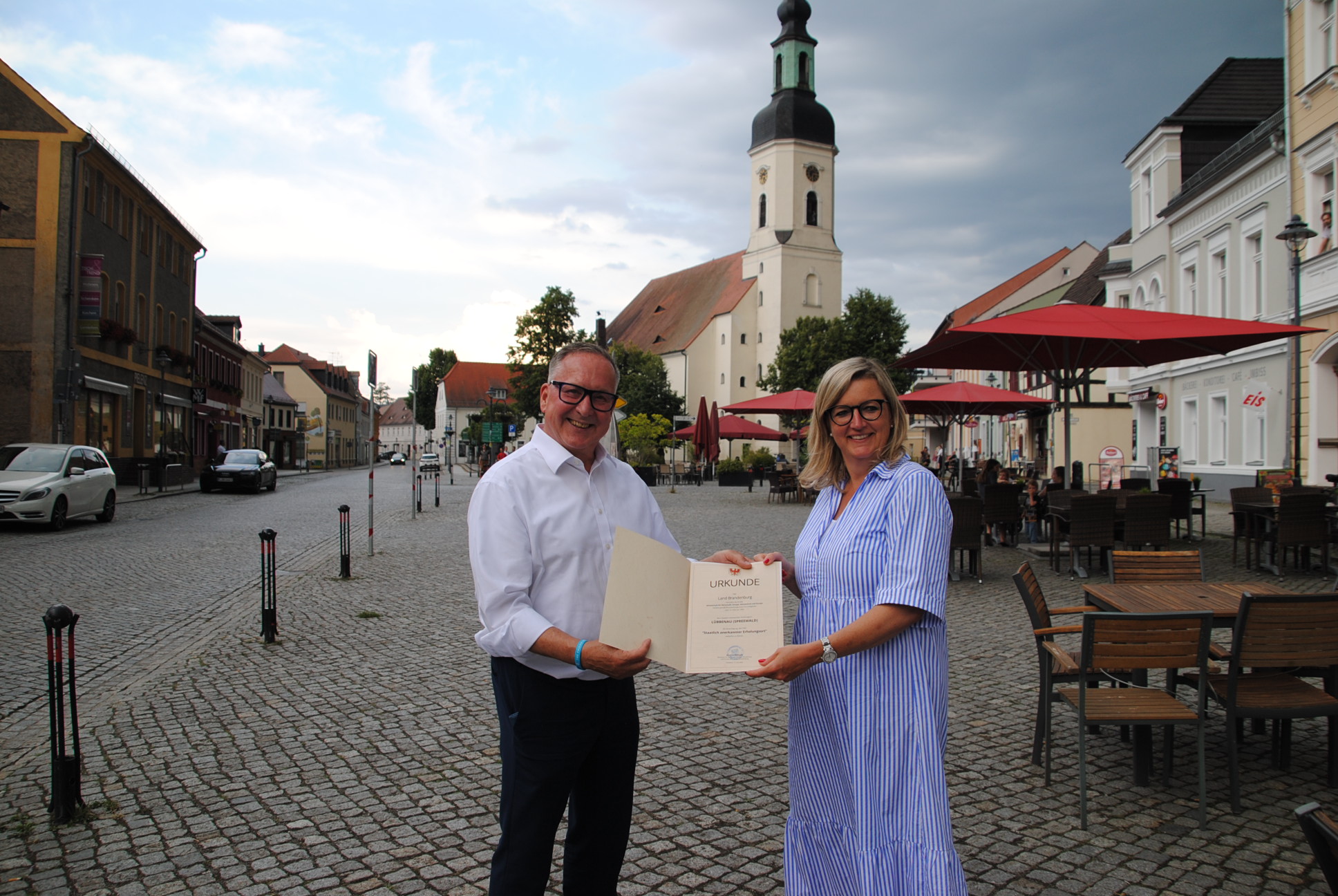 Ministerin Martina Klement überreicht Bürgermeister Helmut Wenzel die Urkunde zur erneuten Anerkennung als staatlich anerkannter Erholungsort Zwei Menschen stehen mit einer Urkunde auf dem Marktplatz in Lübbenau